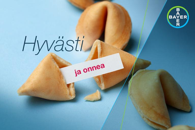 Fortune cookies on a blue surface, with one open revealing a fortune slip. Text on the image reads "Hyvästi ja onnea". A circular logo with "Bayer" is in the corner.