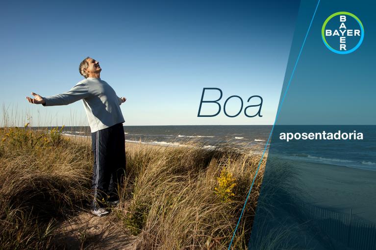 Person standing on a sandy path with arms outstretched near the ocean, under a blue sky. Text reads "Boa aposentadoria." Bayer logo in the corner.