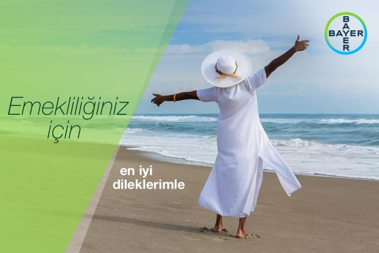 An elderly person in a white dress and hat, arms outstretched, stands on a beach. The text in Turkish conveys retirement best wishes. The Bayer logo is visible.