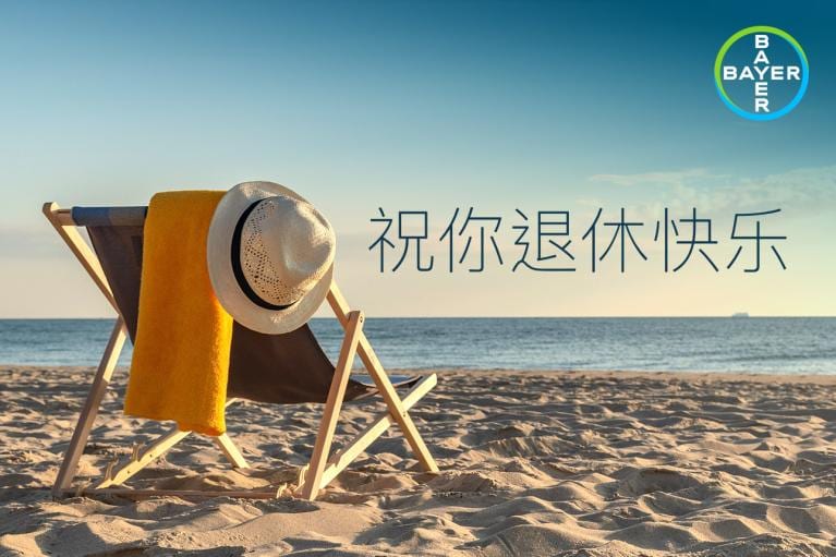 A beach chair with a yellow towel and hat faces the ocean. Bayer logo in the corner. Chinese text reads "祝你退休快乐" meaning "Happy Retirement.
