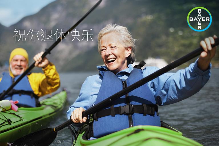 Two elderly people kayaking on a lake, wearing life jackets. Mountains and cloudy sky in the background. Text in Chinese and Bayer logo in the corner.