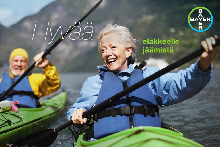 Two older adults kayaking on a lake, wearing life jackets. The man in a yellow jacket and the woman in blue are smiling. Mountains are visible in the background. Finnish text on the image.