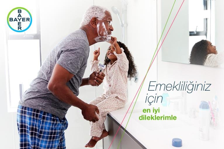 An older man shaving while a child in pajamas playfully watches in a bathroom. Bayer logo and Turkish text are visible.