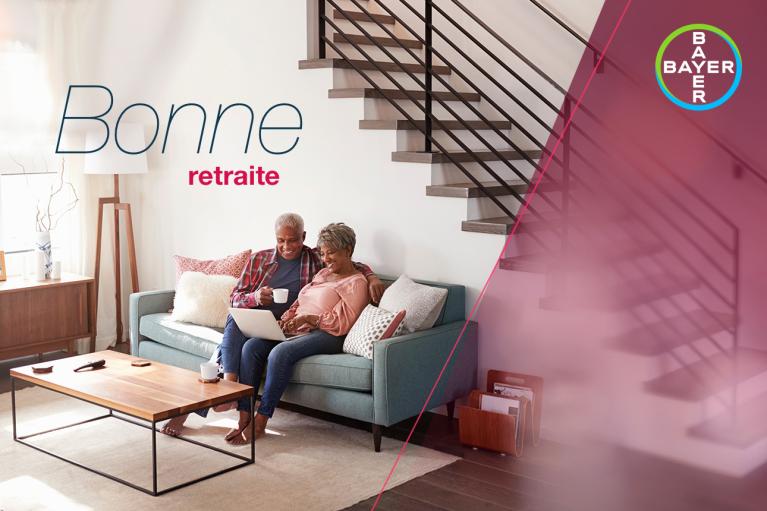 An elderly couple sits on a couch with a laptop and coffee under a staircase. Text reads "Bonne retraite" and includes a Bayer logo.