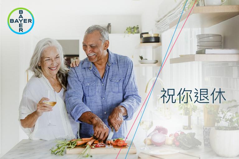 An elderly couple smiling in a modern kitchen, with the man chopping vegetables. A text reads "祝你退休" and the Bayer logo is in the top left corner.