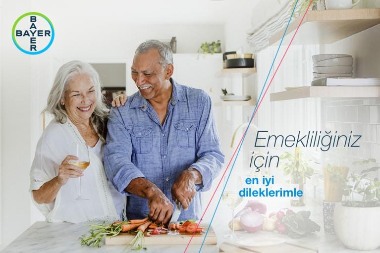 An older couple smiles while preparing vegetables in a bright kitchen. The woman holds a wine glass, and the man stands beside her. Bayer logo and Turkish text are visible on the left.