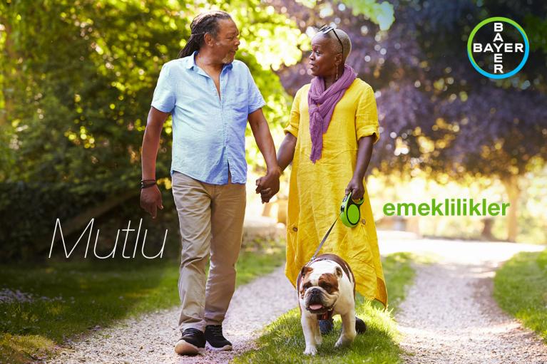 A couple walks hand-in-hand with a bulldog on a sunny path. The words "Mutlu emeklilikler" and a Bayer logo are visible.