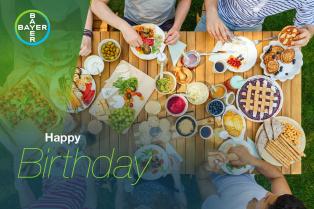 Aerial view of a table with various foods, including pie and fruit, surrounded by people. "Happy Birthday" is written in the bottom left corner next to a Bayer logo.