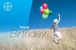 Child holding colorful balloons in a grassy field under a clear blue sky with "Happy Birthday" text and a Bayer logo in the corner.
