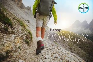 Person hiking on a rocky mountain trail wearing a backpack. Text on image reads "Happy holidays." Bayer logo is in the top right corner.