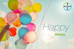 A hand holds colorful balloons against a blue sky with clouds, accompanied by the text "Happy Birthday" and a Bayer logo in the corner.