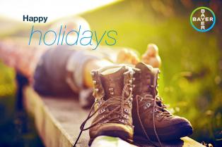 Close-up of hiking boots with a person lying down in the background. Text reads "Happy Holidays" with a Bayer logo in the corner.