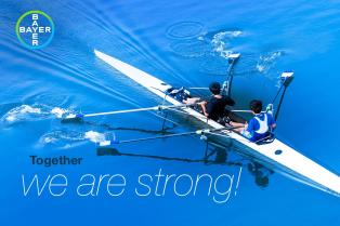 Two people rowing a blue double scull boat on calm water, with the text "Together we are strong!" and a Bayer logo in the corner.