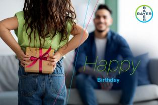 Child hiding a wrapped gift behind their back, while an adult sits in front smiling. Bayer logo is visible in the corner with "Happy Birthday" text on the image.