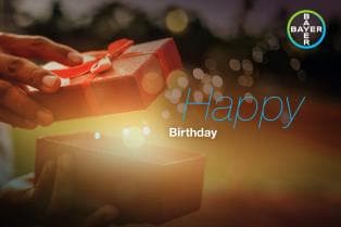 Hands opening a red gift box with sparkles emanating, set against a blurred background. Text reads "Happy Birthday" with a Bayer logo in the top right corner.