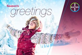 Person dressed in winter clothing with arms outstretched, standing in a snowy landscape. Text reads "Season's greetings" with a Bayer logo in the corner.
