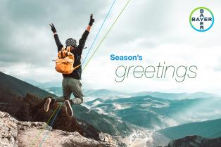 Person jumps on a mountain summit with arms raised. Text reads "Season's greetings" alongside a Bayer logo. Overcast sky and valleys in the background.
