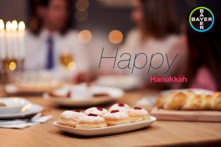 A table with jelly donuts and challah bread, with people in the background. "Happy Hanukkah" text is visible.