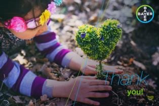 Child planting a heart-shaped shrub in soil while wearing a flower crown, with "Thank you!" text and Bayer logo in the corner.