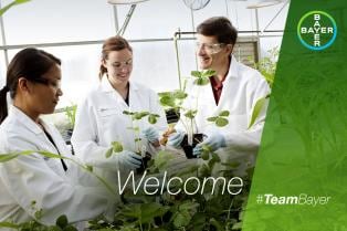 Three people in lab coats and goggles smile while examining plants in a greenhouse. The word "Welcome" is overlaid with the hashtag #TeamBayer in the corner.