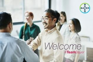 A group of people in an office setting, with one person in the foreground smiling and shaking hands. Logo in the top right corner reads "Bayer." Text states "Welcome #TeamBayer.