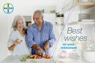 Elderly couple in a kitchen. The man is slicing vegetables, and the woman holds a glass. Text reads, "Best wishes on your retirement." Bayer logo present.