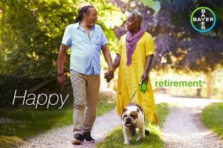 Older African American couple walking a bulldog on a sunny path, with text "Happy Retirement" and Bayer logo.